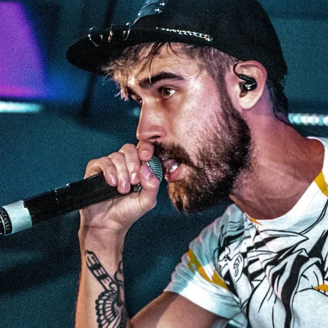 None Like Joshua close-up — teal stage lighting, rapping into the mic with intense focus, anime graphic tee and snapback