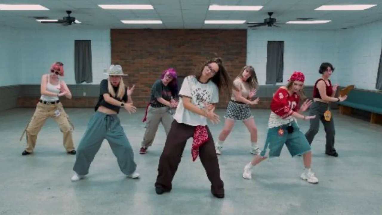 Seven Knox Kpop dancers hit a pose in formation on the floor of the Karns Community Club Event Center, a hall with a brick wall, block walls, and ceiling fans.